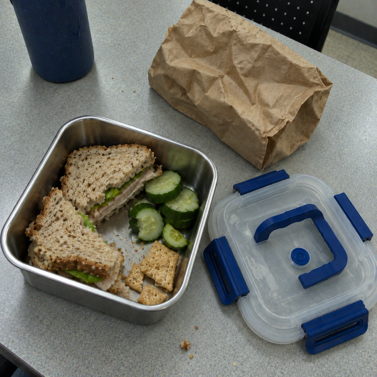 A homemade sandwich, cucumber and crackers in a steel container on a break-room lunch table