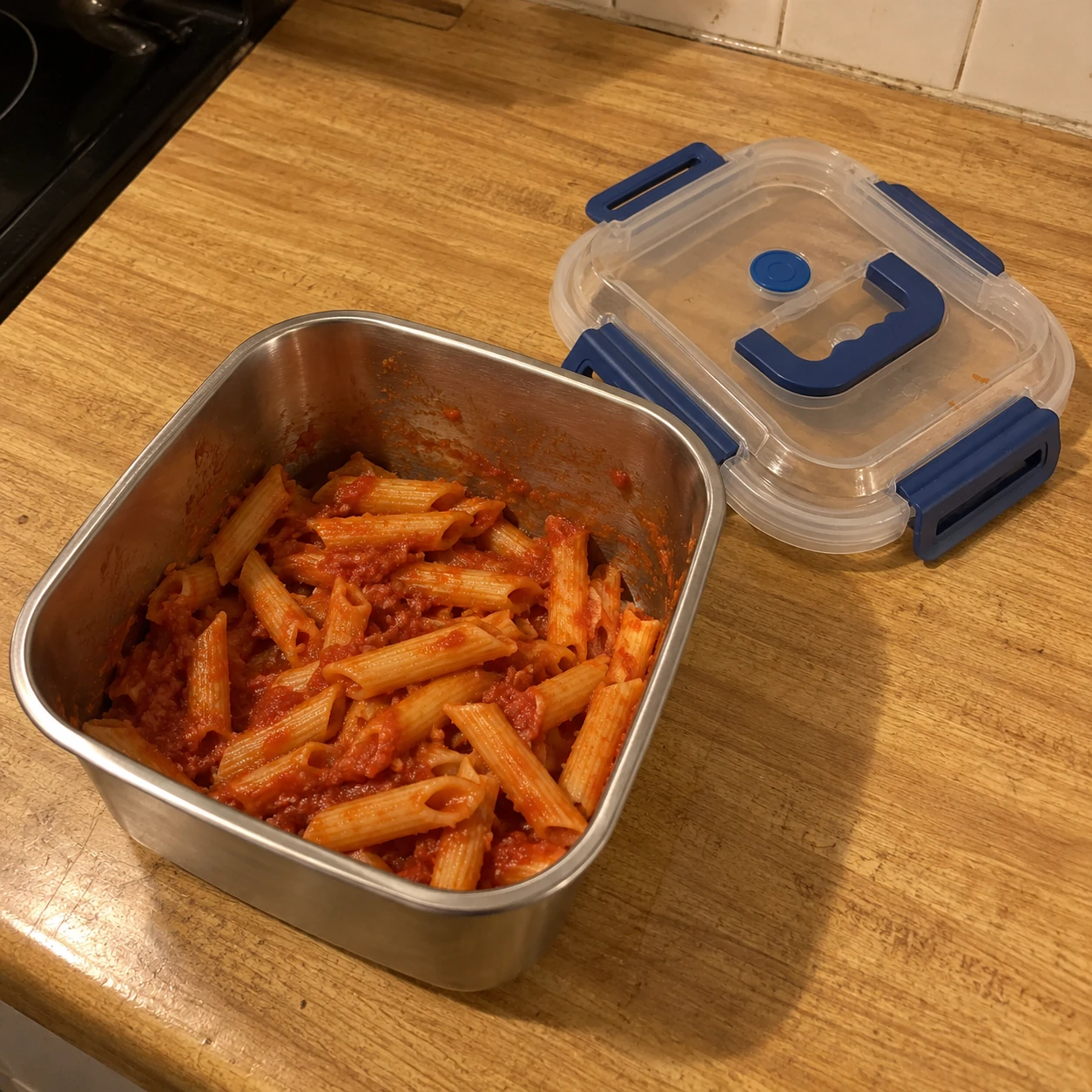 Pasta leftovers in a steel container on a wooden kitchen counter under warm evening light